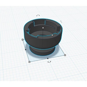 Puede incluir: Objeto cilíndrico negro, renderizado en 3D, con una base redondeada y una sección superior más ancha. La parte superior tiene un interior empotrado con cortes angulares. El objeto se encuentra sobre un fondo de cuadrícula, lo que sugiere un diseño o modelo digital.
