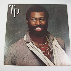 May include: A square vinyl record cover featuring a portrait of a man with a beard, wearing a grey collared shirt and a light-colored scarf. He wears multiple gold chains around his neck. The letters Tp are printed in white in the top left corner.