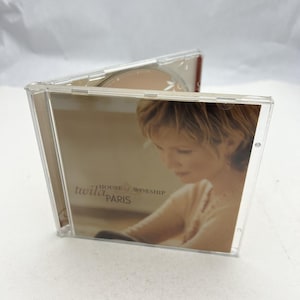 May include: A clear plastic CD case, open to reveal the disc and album art. The album art features a woman with short blonde hair and the text "twila paris HOUSE of WORSHIP PARIS". The CD is visible inside the case.