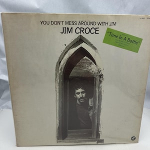 Puede incluir: Una portada de álbum de vinilo con una foto en blanco y negro de Jim Croce mirando por una ventana. El título del álbum es "You Don't Mess Around With Jim Jim Croce" y el título de la canción es "Time In A Bottle".