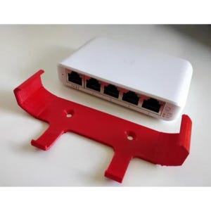 Puede incluir: Un conmutador de red blanco con múltiples puertos y un soporte de montaje rojo brillante. El soporte tiene una forma única con dos agujeros y está diseñado para sujetar el conmutador de forma segura. Los dispositivos están sobre una superficie blanca.