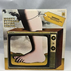 May include: A vintage vinyl record cover featuring a giant foot stepping on a television screen. The cover art is for the album "Monty Python's Flying Circus" and includes the text "Warning! This album is original material from the TV series".