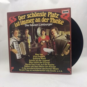 May include: A vintage vinyl record album cover featuring a group of people playing accordions in a bar setting. The album cover is titled "Der schönste Platz ist immer an der Theke" and features the text "Die fidelen Limburger" and lyrics about beer, Spain, and a beautiful day. The album cover is in a red, white, and black color scheme.