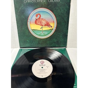 Puede incluir: Una portada de álbum de vinilo con un flamenco rosa de pie en un cuerpo de agua. La portada del álbum es verde y tiene el texto "Christopher Cross" impreso en blanco. El disco es negro y tiene una etiqueta blanca con el texto "Christopher Cross" y el título del álbum "Christopher Cross" impreso en él.