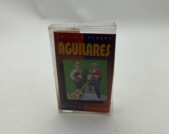 以下が含まれることがあります： エミリオとジェナロのアルバム「Aguilares」が写った透明なカセットテープケース。カバーアートには、ギターとアコーディオンを持った伝統的な衣装の男性が2人描かれています。「Me Estoy Acostumbrando」の文字も確認できます。