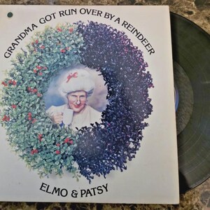 May include: Album cover for "Grandma Got Run Over by a Reindeer" by Elmo & Patsy. The cover features a portrait of a woman in a wreath, holding a mug. The vinyl record is visible on the right side.