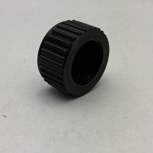 May include: A black, cylindrical object with a ridged exterior and a threaded interior. The object appears to be made of a solid material and is set against a plain, light-colored background. The ridges run parallel around the circumference.