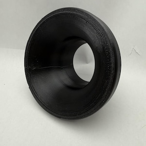 May include: A black, 3D-printed funnel with a wide, flared opening and a circular hole. The funnel has a textured surface and appears to be made of plastic. The object is designed to direct liquids or solids into a narrow space.