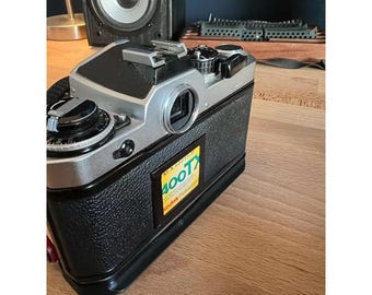 May include: A vintage silver and black film camera with a black leather grip. The camera has a lens, dials, and a flash attachment. A yellow and green sticker on the side reads "RTX 400T". The camera is on a wooden surface.