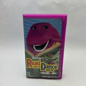 May include: A purple VHS tape of "Barney's Read with Me Dance with Me." The cover features Barney the dinosaur reading a book, with the title in yellow and blue. The tape is in a pink plastic case and says "52 Minutes" and "Never Seen On TV."