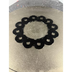 Puede incluir: Un objeto metálico negro con forma de flor, con una abertura circular central y formas de pétalos espaciadas. Cada pétalo tiene un pequeño agujero circular. El objeto está sobre una superficie texturizada de color plateado.