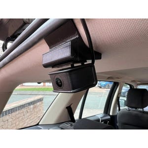 May include: A black dash camera mounted inside a vehicle. The camera is attached to the ceiling and has a lens facing forward. The interior of the car is visible, including the seats and windows. The camera appears to be a 3D-printed design.