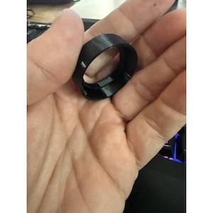 May include: A black, 3D-printed ring with a rectangular cross-section. The ring is held in a person's hand, with the focus on the ring's shape and texture. The ring appears to be made of plastic or a similar material.