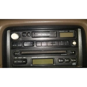 May include: A black Toyota car stereo head unit featuring a cassette player labeled Logic Control Deck, a CD player slot, two digital displays, and various control buttons including volume, tuning, track selection, and radio band switches.