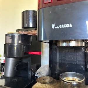 May include: A black and brown espresso machine with the brand name "easy GAGGIA" visible. The machine has a grinder, a portafilter, and a water reservoir. A red lever and various buttons are also visible.