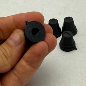May include: A set of four small black plastic control knobs with a textured finish. Each knob features a circular top with a D-shaped center hole for attachment and a small pointed indicator mark on the outer edge for precise positioning.