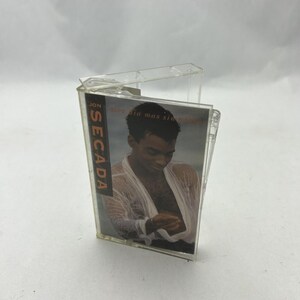 May include: A clear cassette tape case holding a music album. The album cover features a man in a wet, white shirt. The album title is "Secada" with the artist's name "Jon" visible on the spine.