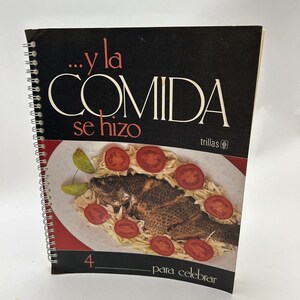 Könnte beinhalten: Ein spiralgebundenes Kochbuch mit schwarzem Einband und dem Titel "...y la COMIDA se hizo" in weißer Schrift. Der Einband zeigt ein Foto von einem gebratenen Fisch mit Tomaten und Kohl auf einem weißen Teller. Das Buch ist Teil der Reihe "Trillas" und ist Band 4. Der Text "para celebrar" befindet sich am unteren Rand des Einbands.