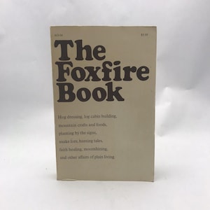 May include: A vintage book titled "The Foxfire Book" with brown lettering on a beige cover. The book's subtitle lists topics such as hog dressing, log cabin building, and moonshining. The book is standing upright.