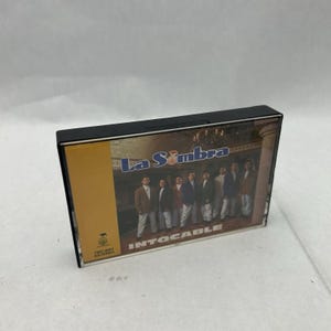 May include: A black cassette tape case featuring the album cover for "La Sombra" by Intocable. The album cover shows a group of men in suits. The cassette case has a yellow label with the album title and a logo.