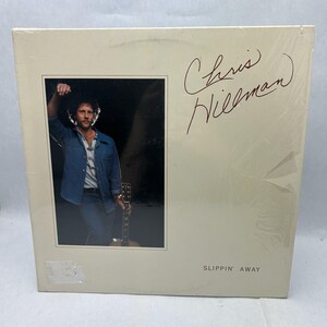 Peut inclure: Un album vinyle vintage intitulé "Slippin' Away" de Chris Hillman. La couverture de l'album présente une photographie d'un homme en veste en jean tenant une guitare. La signature de l'artiste est visible dans le coin supérieur droit.