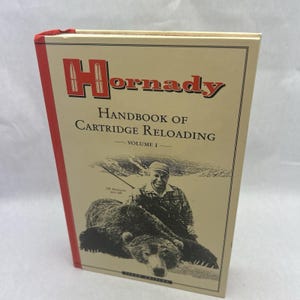 May include: A hardcover book titled "Hornady Handbook of Cartridge Reloading, Volume I." The cover features a man with a bear and the Hornady logo in red. The book has a red spine and a cream-colored background.