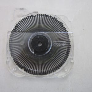 以下が含まれることがあります： 黒い書体の透明なプラスチック製タイプライターホイール。ホイールは透明なプラスチックケースに収められています。ホイールには「brother」の文字が印刷されています。ホイールは、小さな黒い文字が多数ある円形デザインです。