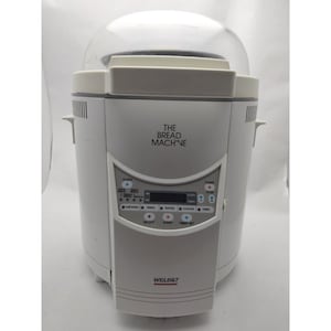 Könnte beinhalten: Eine weiße Welbilt Brotbackmaschine mit einem transparenten Kunststoffdeckel. Die Maschine verfügt über ein digitales Display und Bedientasten zum Kneten, Gehen, Backen und Abkühlen. Der Text "THE BREAD MACHINE" ist auf der Vorderseite aufgedruckt.