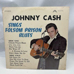 May include: A vintage vinyl record album cover featuring Johnny Cash. The cover is white with black text that reads "Johnny Cash Sings Folsom Prison Blues". The image shows Johnny Cash playing a guitar with a microphone in front of him. The album cover has a black and white photo of Johnny Cash.