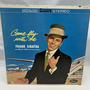 May include: Vintage vinyl record album cover for Frank Sinatra's "Come Fly With Me". The cover shows Sinatra in a suit and fedora against a blue sky with airplanes. The album is labelled "STEREO" and "Capitol Full Dimensional".