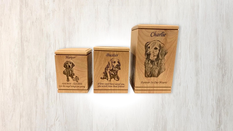 May include: Three wooden pet urns with engraved images of dogs and personalized text. The urns are in different sizes and feature the names "Harper", "Buster", and "Charlie".