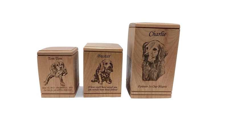 May include: Three wooden pet urns with engraved images of dogs and personalized text. The urns are different sizes and feature the names "Tom Tom", "Buster", and "Charlie".