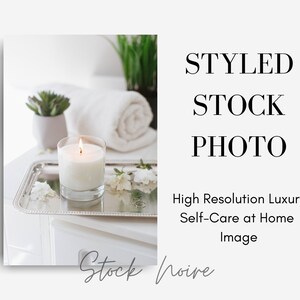 May include: A white candle burning in a glass jar on a silver tray with white flowers and a white towel. The tray is on a white surface.