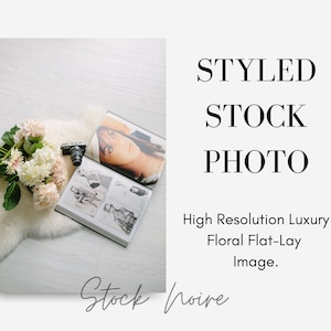 May include: A flat lay styled stock photo featuring a bouquet of white and pink hydrangeas, a vintage camera, and an open magazine with a fashion model on the cover. The image is set on a white faux fur rug and a white wood background.