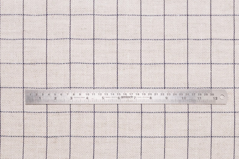 May include: A close-up of a white fabric with a subtle herringbone pattern and a thin black grid. A ruler is placed on the fabric, showing measurements in inches.
