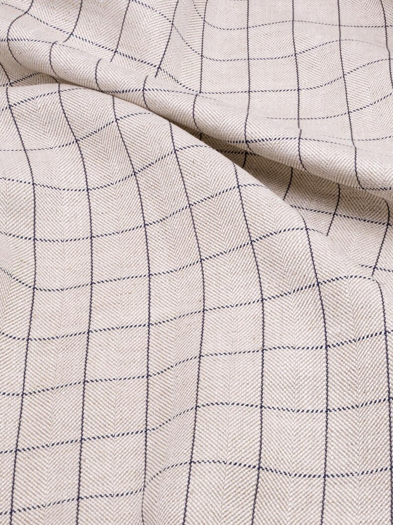 May include: A close-up of a beige fabric with a fine, dark blue checkered pattern. The fabric is woven with a herringbone texture.