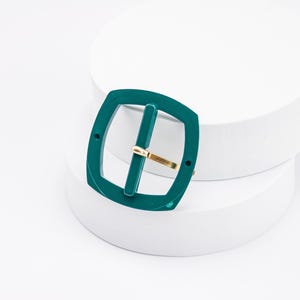 May include: A teal green plastic belt buckle with a gold metal bar. The buckle is on a white surface.