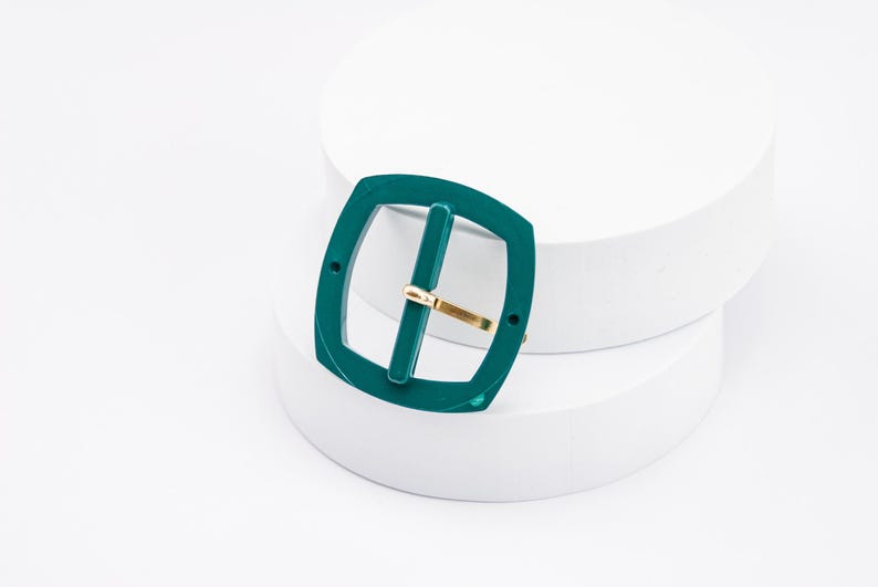 May include: A teal green plastic belt buckle with a gold metal bar. The buckle is on a white surface.