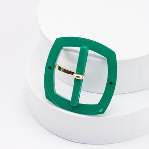 May include: A green plastic belt buckle with a gold metal bar. The buckle is square shaped with rounded corners.