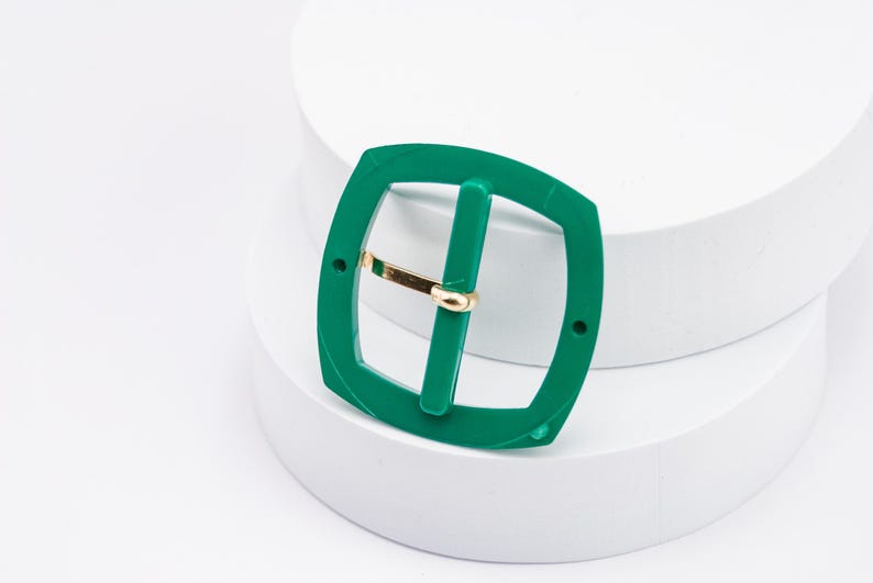 May include: A green plastic belt buckle with a gold metal bar. The buckle is square shaped with rounded corners.