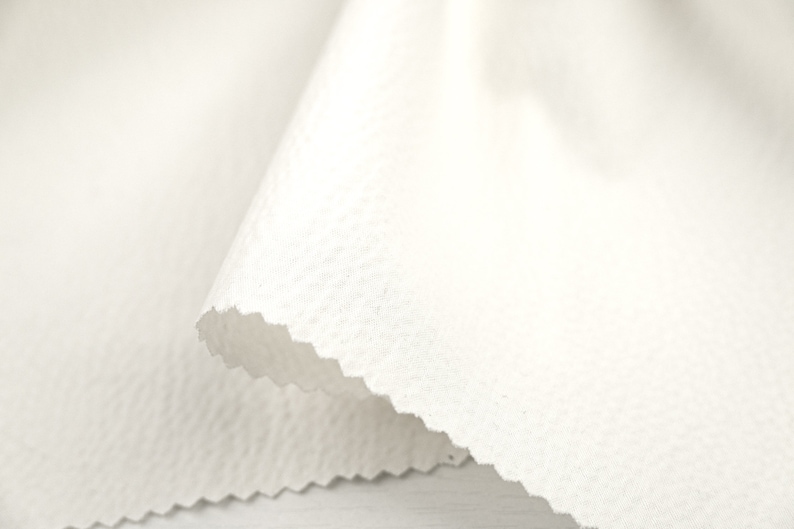 May include: White fabric with a textured surface. The fabric is folded over itself, creating a wave-like shape.