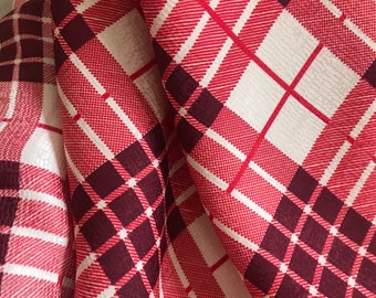 May include: Red and white plaid fabric with a burgundy checkered pattern. The fabric is folded and draped, showing the texture and pattern.