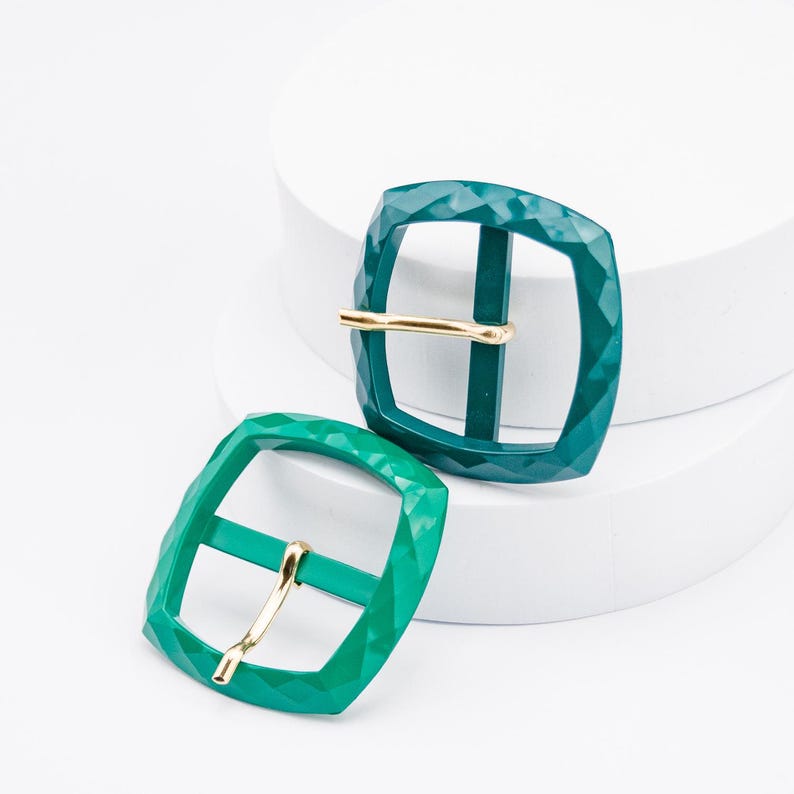 May include: Two green metal belt buckles with a gold metal bar. The buckles have a diamond-shaped pattern.