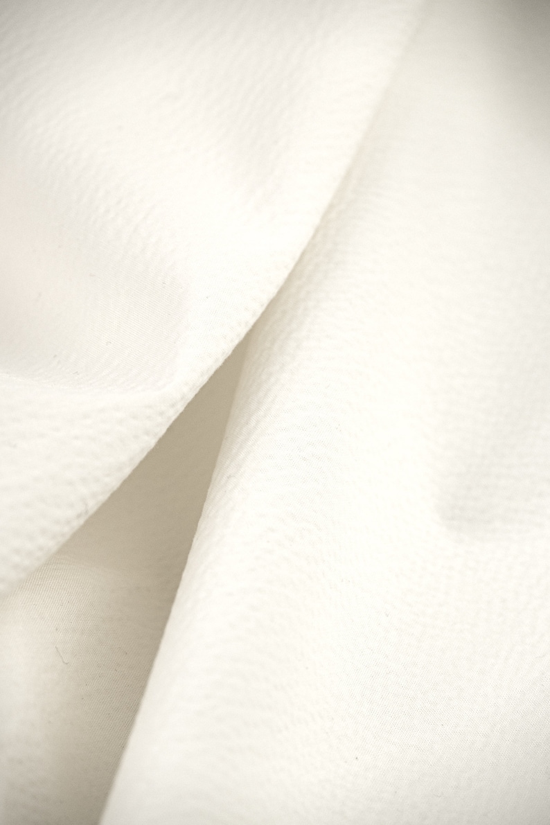 May include: Close-up of a white fabric with a textured surface. The fabric is folded in half, creating a soft, flowing look.