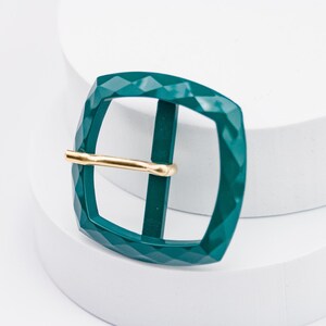 May include: A teal green metal buckle with a gold metal bar. The buckle has a diamond-shaped pattern.