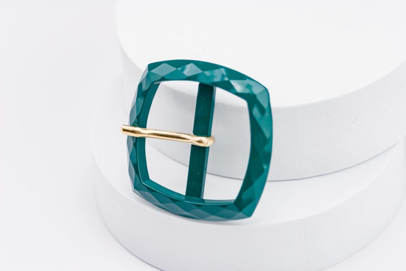 May include: A teal green metal buckle with a gold metal bar. The buckle has a diamond-shaped pattern.