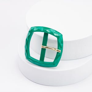 May include: A green metal buckle with a diamond pattern. The buckle is square shaped and has a gold metal pin.