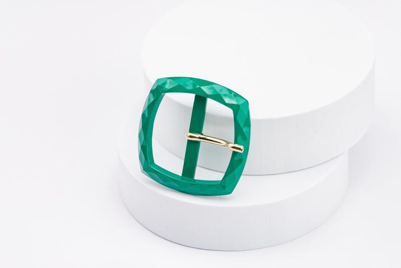 May include: A green metal buckle with a diamond pattern. The buckle is square shaped and has a gold metal pin.
