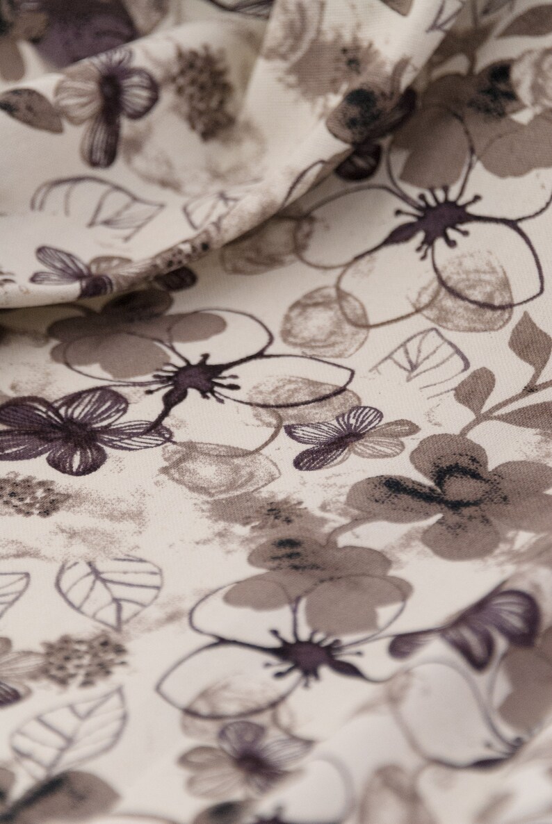 May include: A close-up of a white fabric with a floral pattern. The flowers are brown and black and have a watercolor effect. The fabric is soft and has a subtle texture.