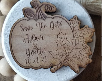 Op de afbeelding: Een houten save-the-date-bord in de vorm van een pompoen met een esdoornblad. Het bord heeft de tekst "Save The Date Adam & Yvette 10.21.21".